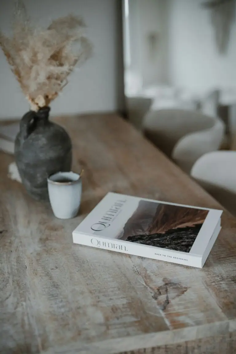 A book on a worn wooden table beside a vase of dried grasses