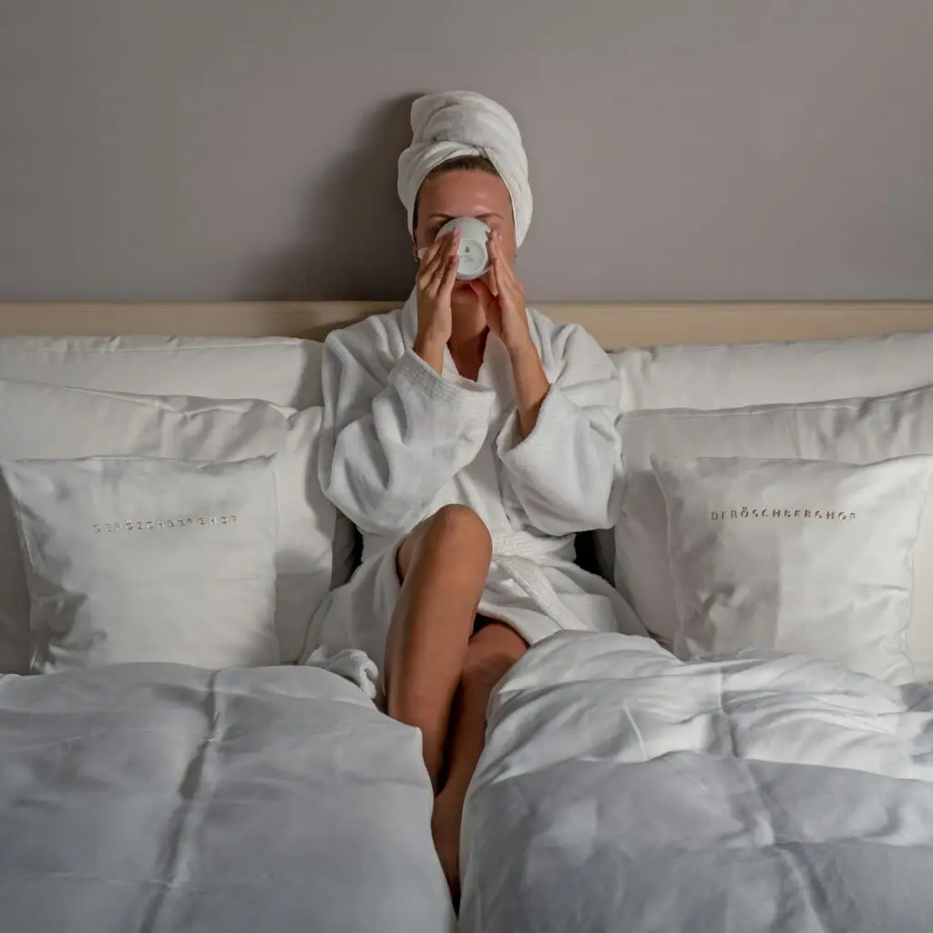 A woman in a white robe and towel turban relaxing on a crisply made hotel bed