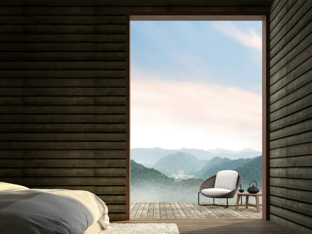 A timber-lined bedroom opening onto a terrace with a single chair and misty mountains beyond