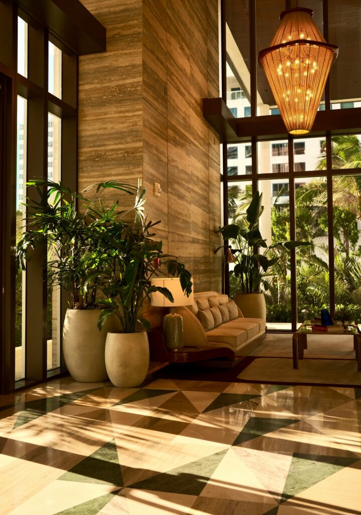 A double-height hotel lobby with a glowing chandelier, tall palms and a patterned stone floor
