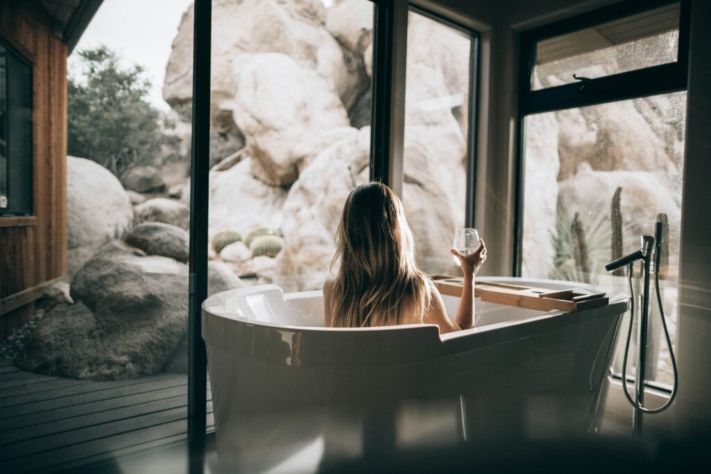 A woman alone in a freestanding bath beside a window onto rocks and sky